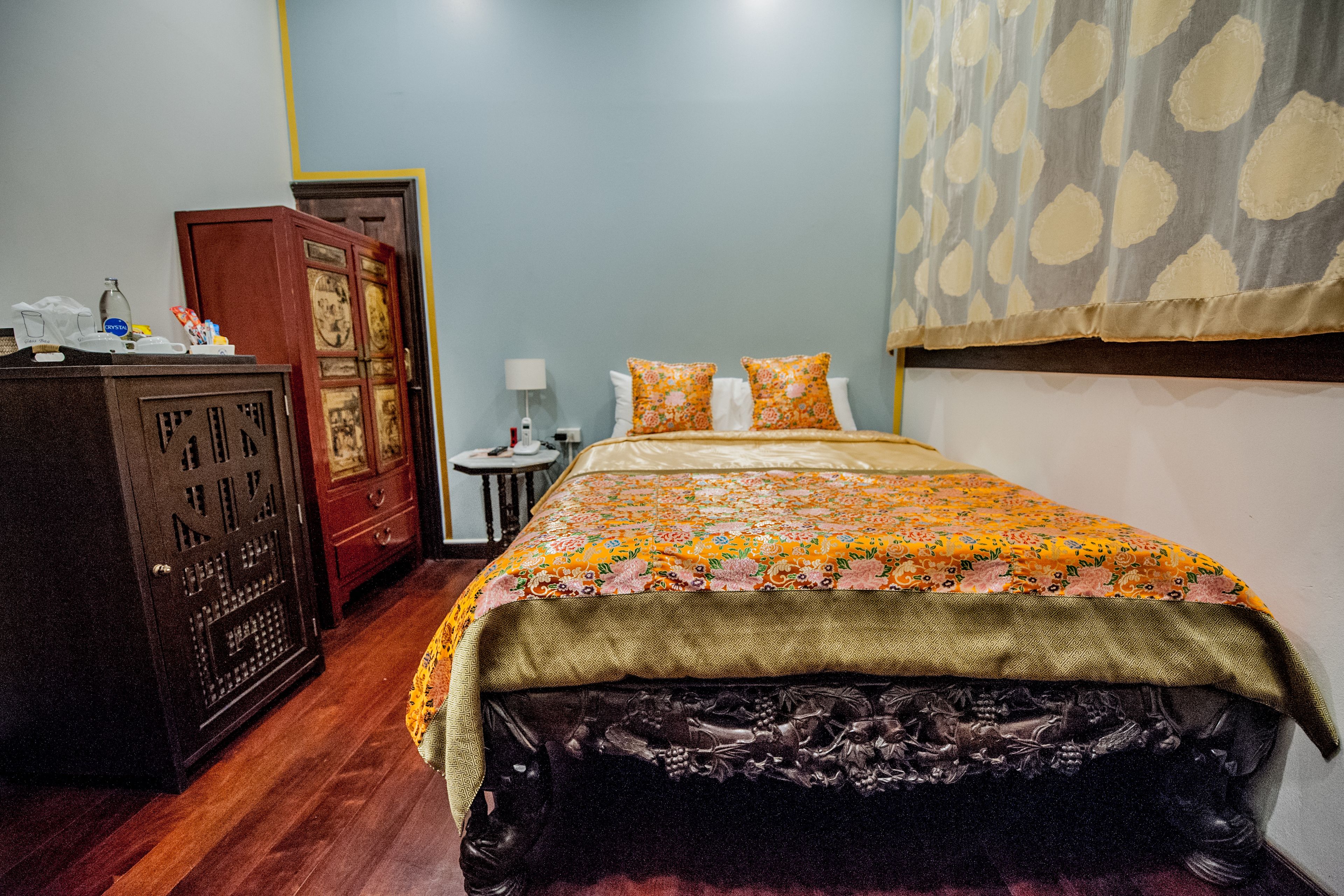 A bedroom with a wooden wardrobe, a small round table with a lamp, and a bed with floral bedding.