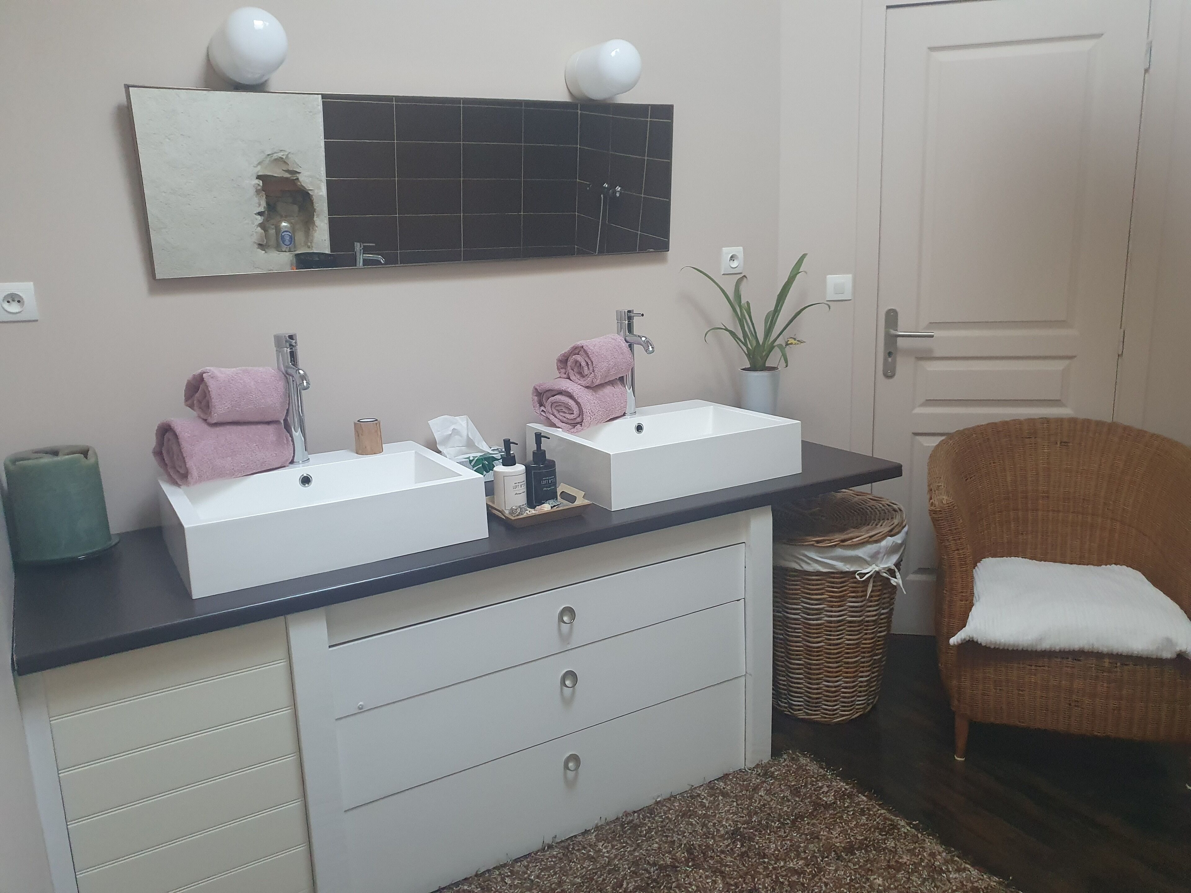 Un baño con dos lavamanos, un espejo, una planta en maceta y una silla de mimbre.
