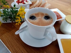 Eine Kaffeetasse mit Milchschaumkunst auf einem Untertasse, eine kleine Schale Erdbeeren, eine Brotteller und ein gelbes Blumenarrangement auf einem Holztisch.
