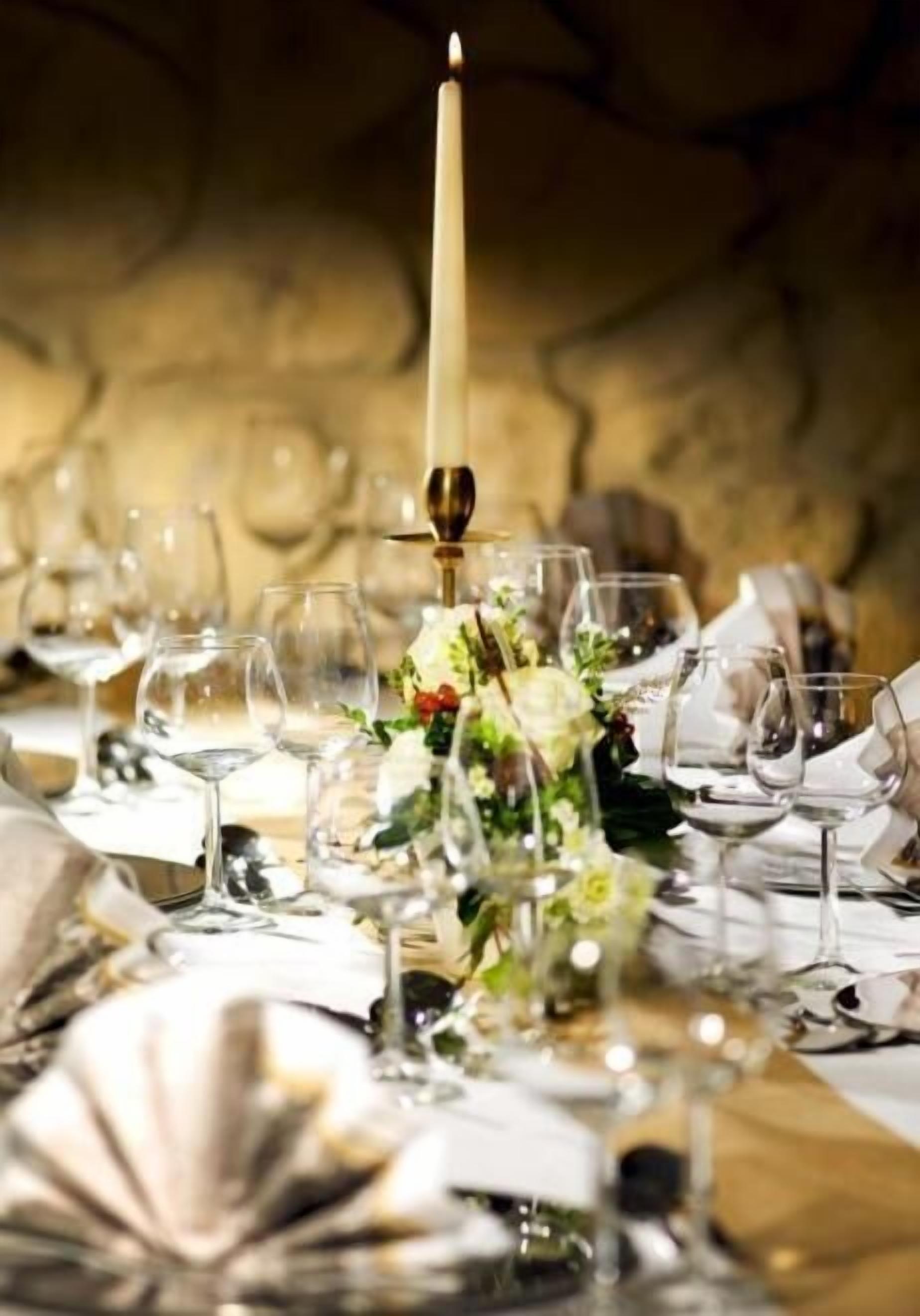 Une table dressée pour un dîner formel, agrémentée d’un centre de table composé de bougies, de verres transparents et d’un mur en pierre en arrière-plan.