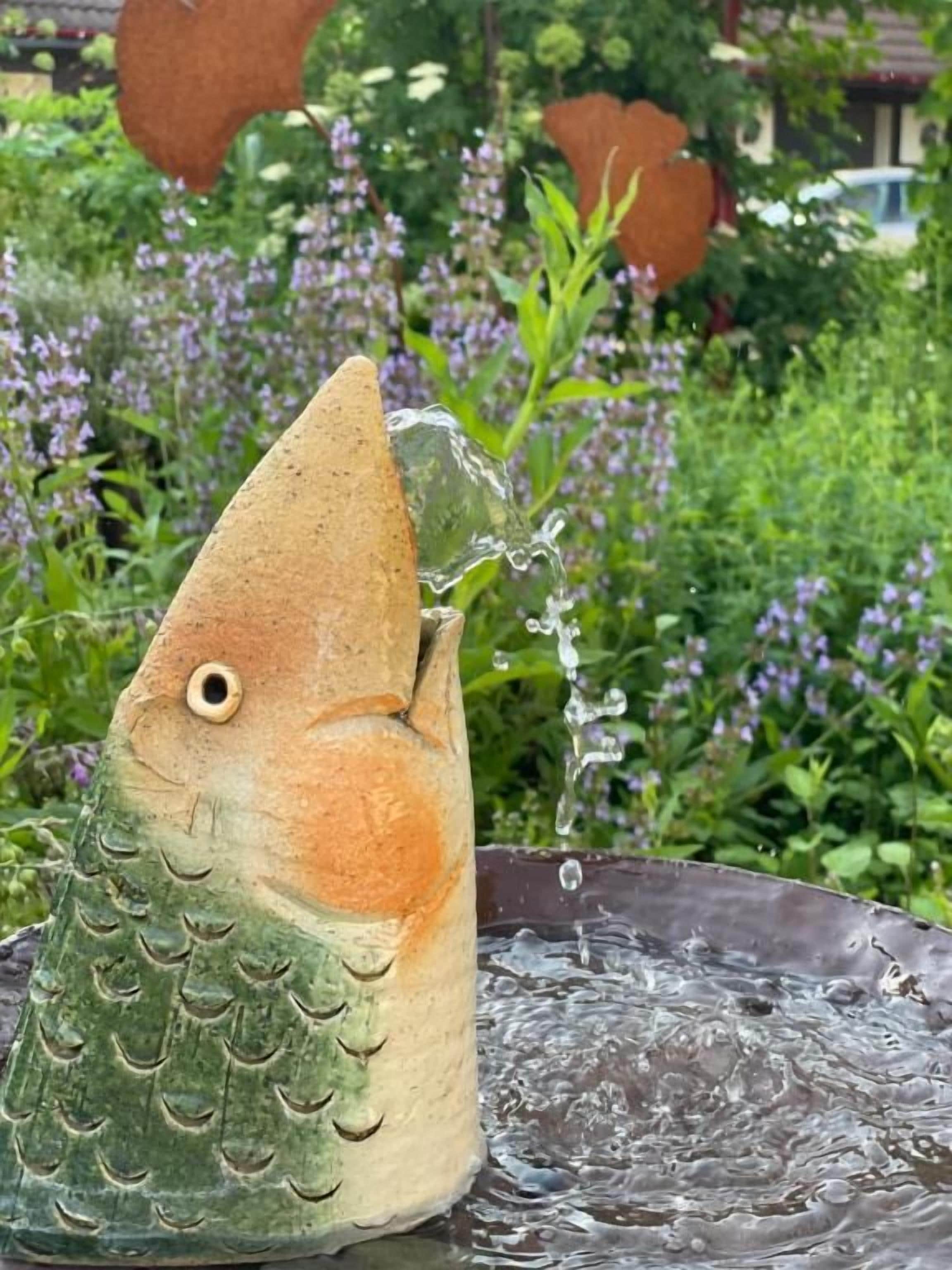 Una fuente con forma de pez, de la cual fluye agua, con un fondo de follaje y flores moradas.