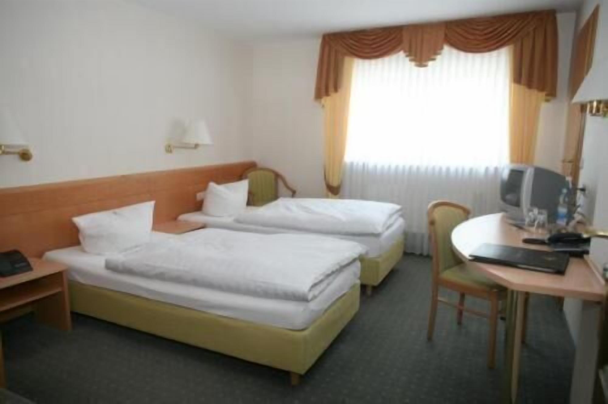 A hotel room with two single beds, a desk with a chair, a television, and a window with curtains.