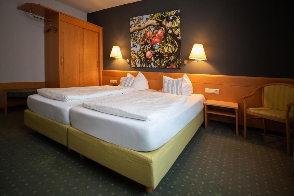 Cama doble con dos almohadas, un armario de madera, una mesita y una silla en una habitación de hotel.