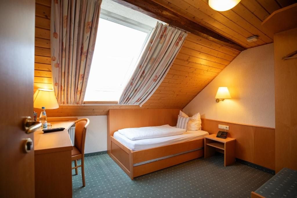 A small, cozy room with a bed, a desk, and a chair. There is a window with curtains, a lamp, and a small bedside table.