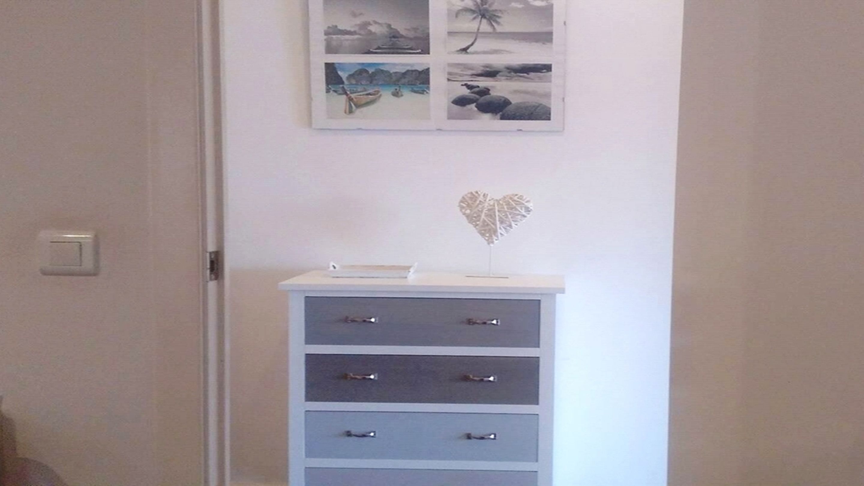 Une pièce avec un mur blanc, une petite armoire blanche avec des tiroirs, et au-dessus, un collage encadré représentant des scènes de plage.