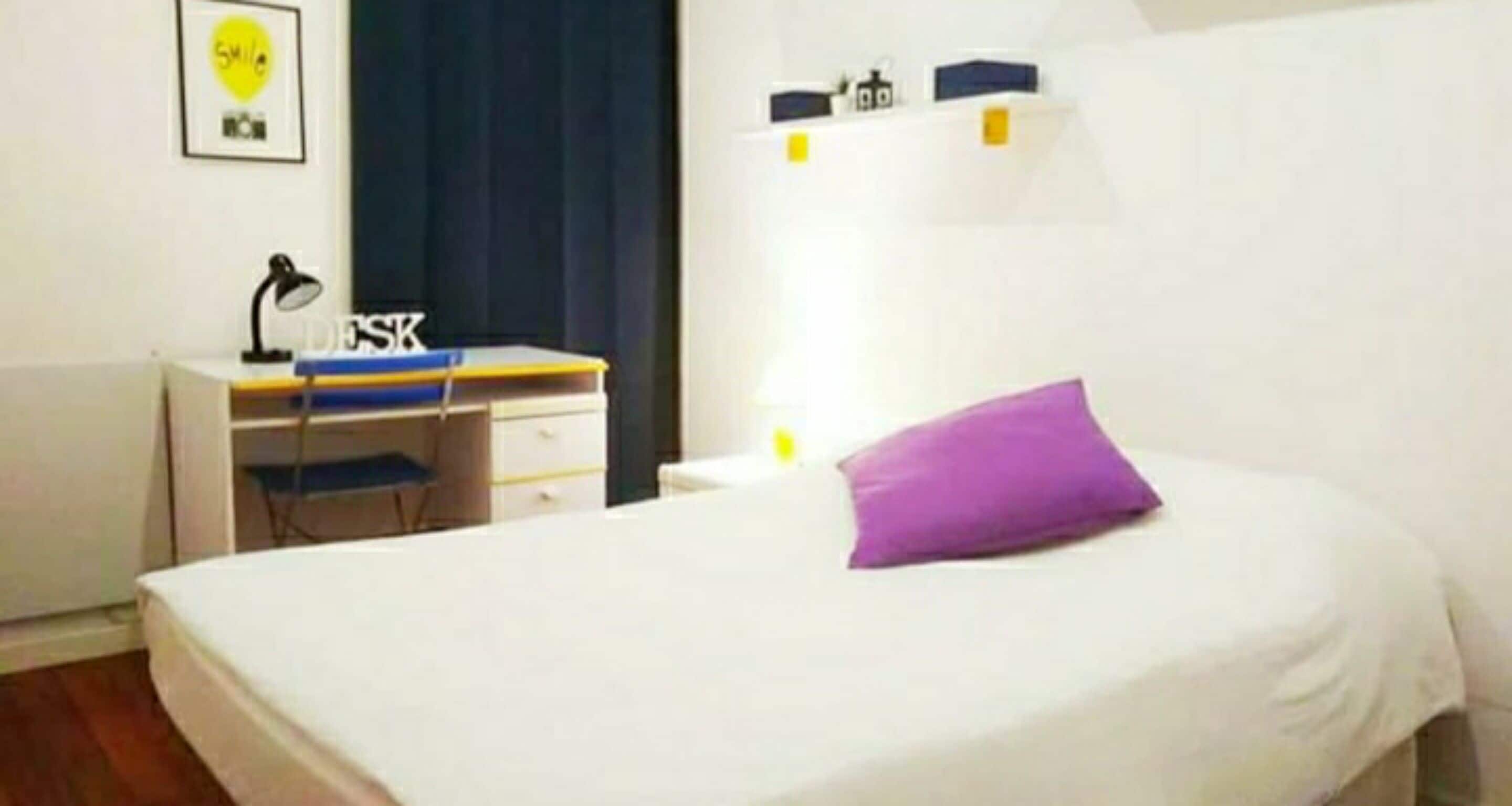 Un dormitorio con una cama, un escritorio con una silla, una lámpara y un estante en la pared con objetos.