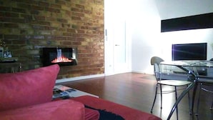 A living room with a brick fireplace, a red sofa, a glass table with chairs, and a flat-screen TV mounted on the wall. - Habitacions Sant Pere Claver (Tarrega)