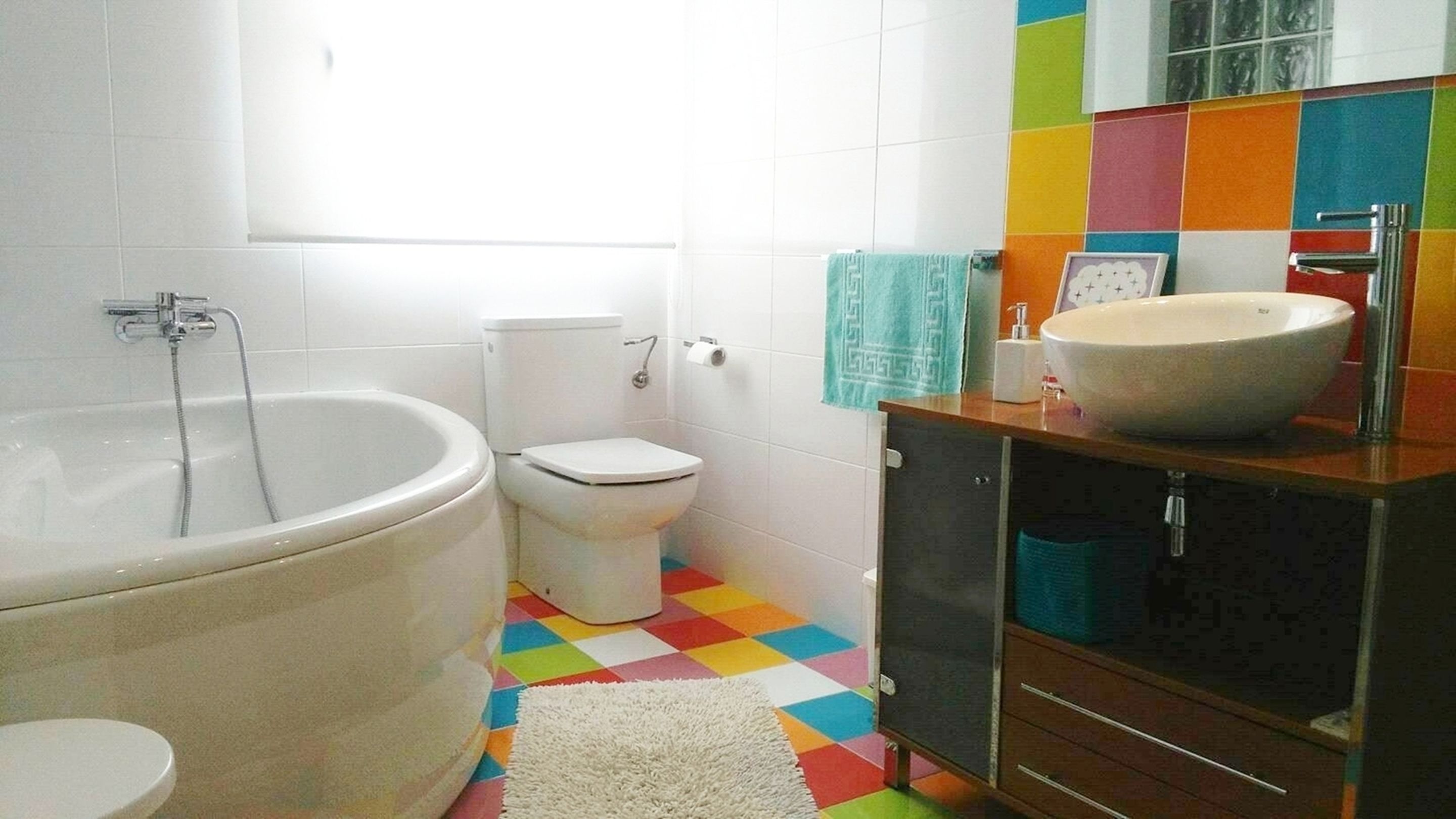 Un baño con bañera, inodoro y lavabo con espejo. El suelo presenta un estampado de cuadros de colores.