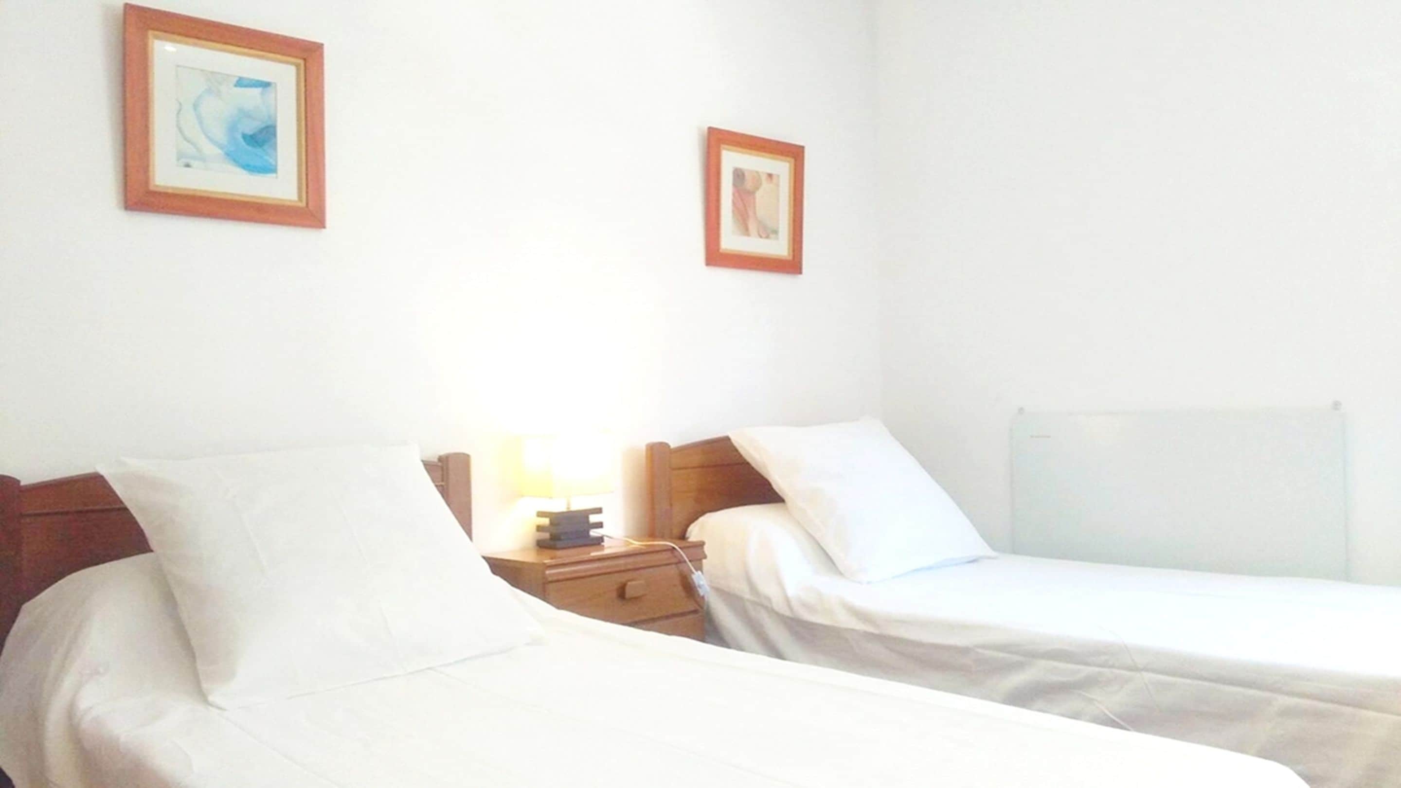 Een hotelkamer met twee éénpersoonsbedden, een nachtkastje met een lamp en twee ingelijste foto's aan de muur.