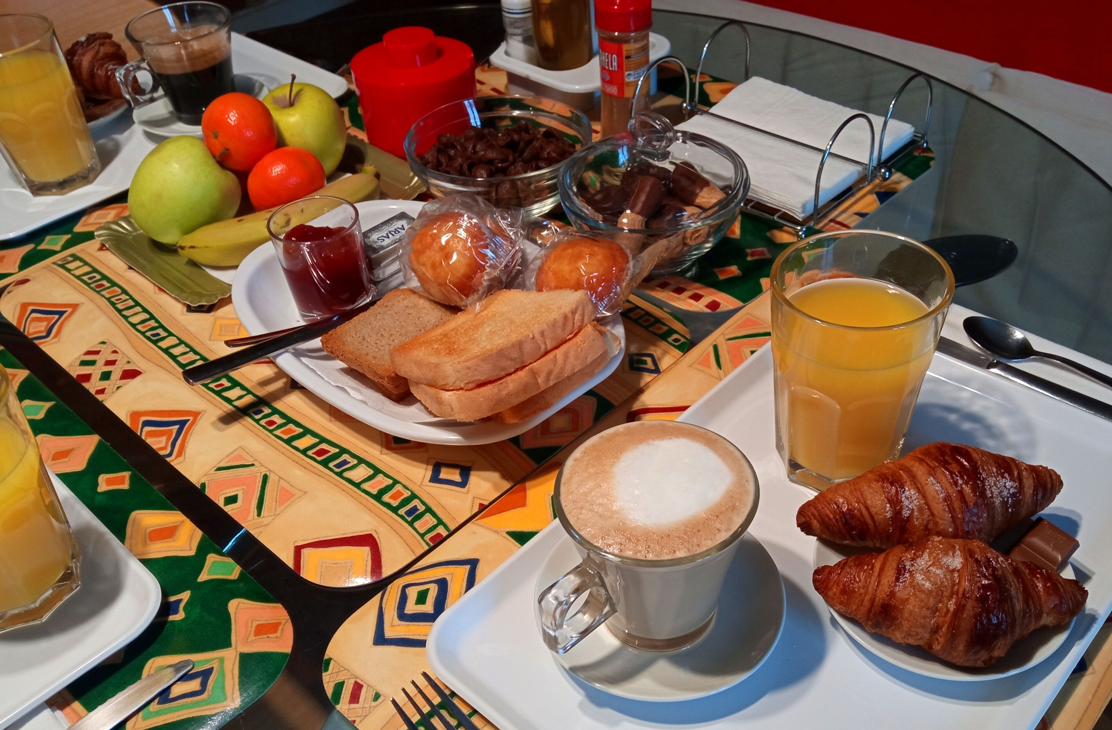 Una mesa de desayuno con diversos alimentos y bebidas, como croissants, tostadas, fruta y una taza de café.