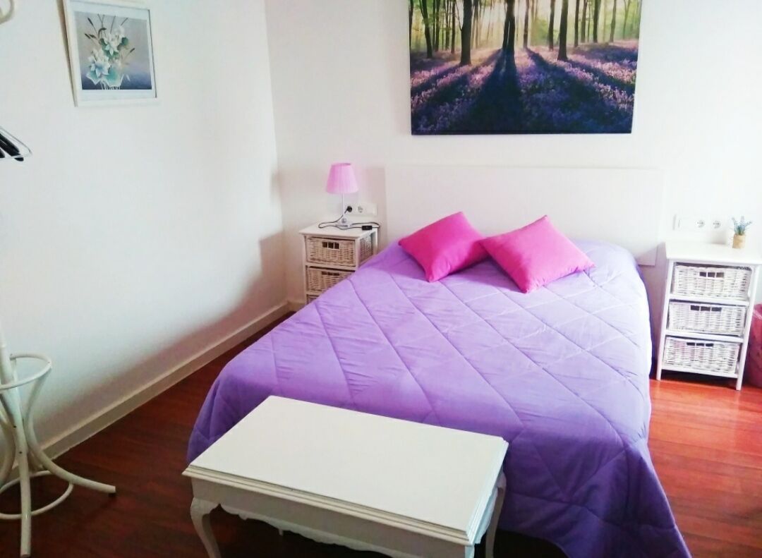A bedroom with a purple bedspread, two pink pillows, a white bedside table, a wicker nightstand, and a painting of a forest path on the wall.