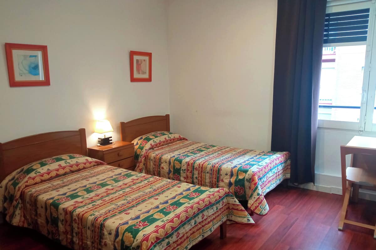 Ein Zimmer mit zwei Holzbetten, floralem Bettzeug, einem Nachttisch mit Lampe und einem Fenster mit Vorhängen.