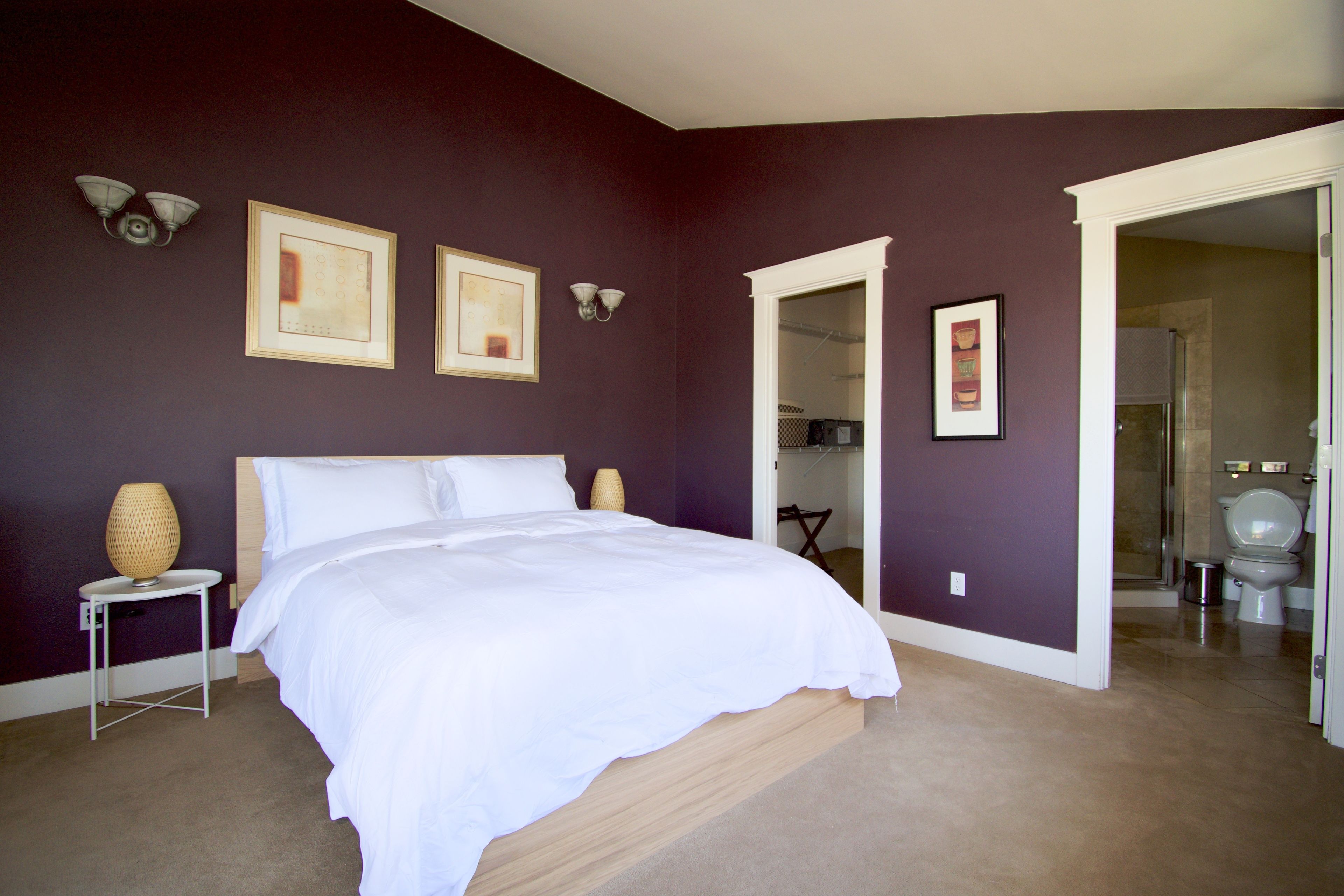 A bedroom with a large bed, a nightstand, and a wall-mounted lamp. There are framed pictures on the wall and a door leading to another room.