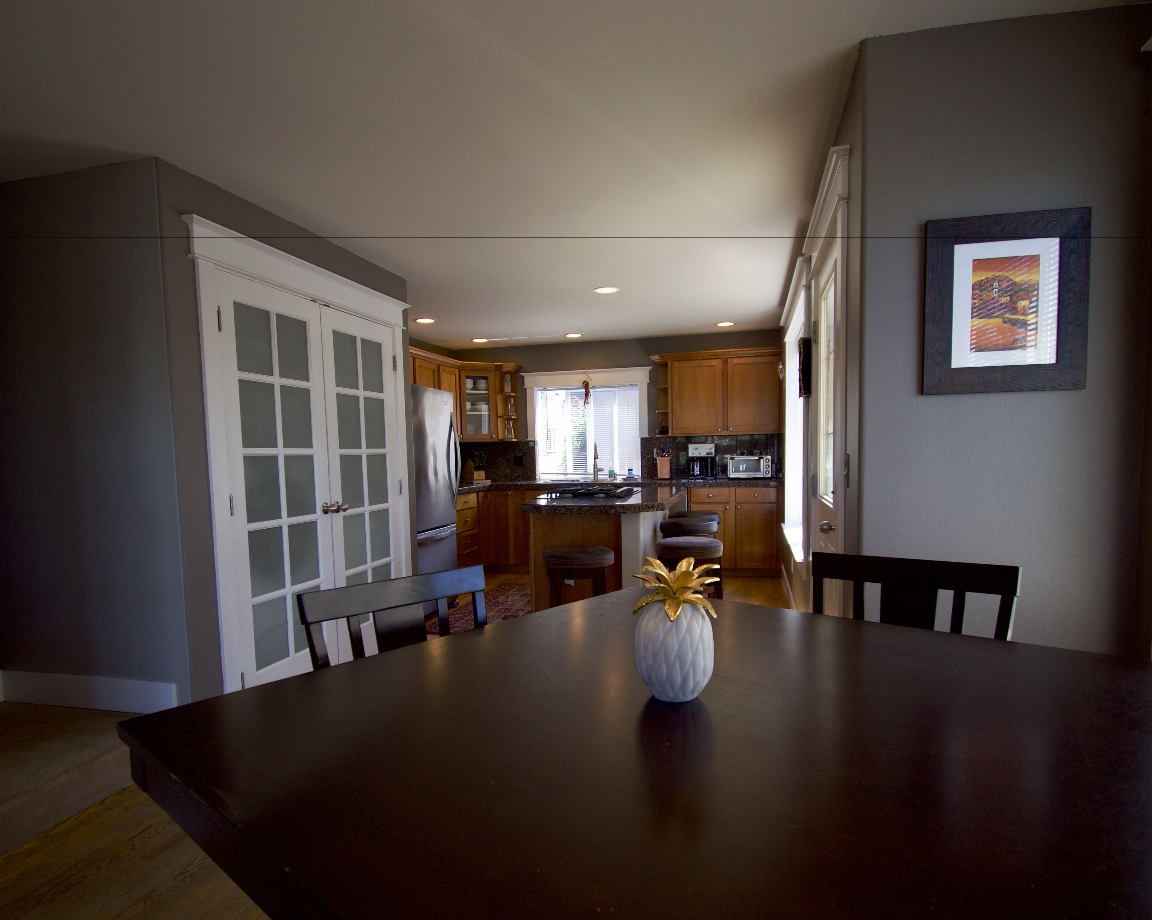 A dining area with a dark wooden table, a vase, and chairs. A kitchen with wooden cabinets and a window is visible in the background.