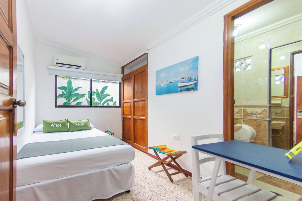 Un dormitorio con cama, escritorio y silla. Hay una ventana con vistas a plantas y un cuadro con barcos en la pared.