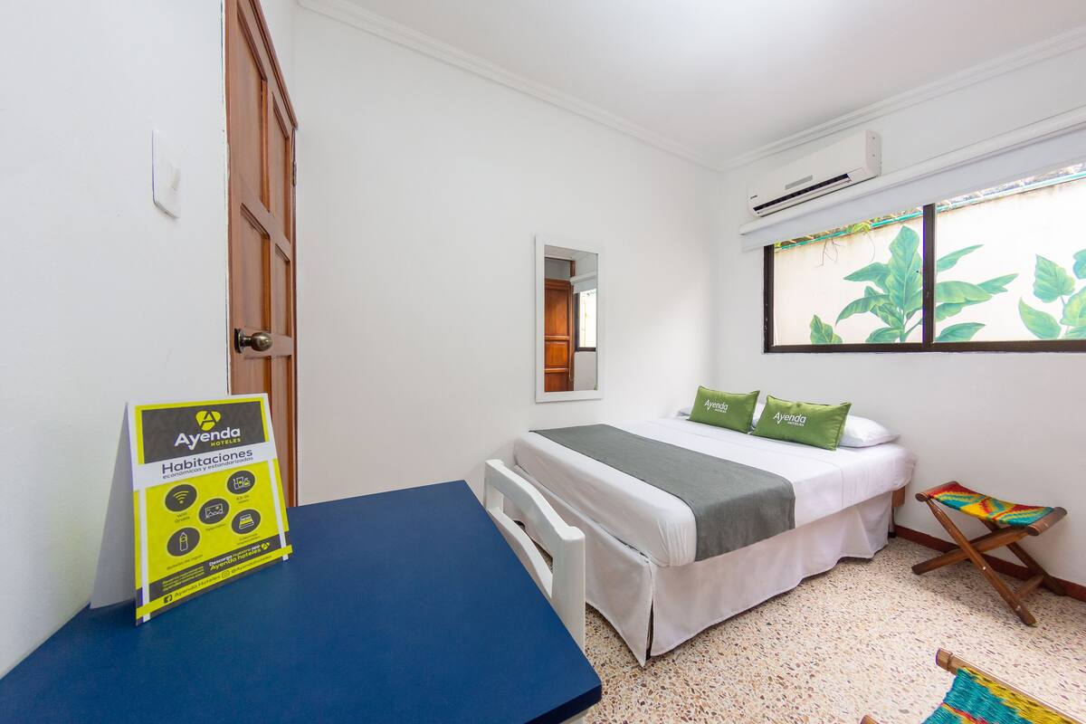 Una habitación de hotel con una cama, una mesita, una silla, un espejo y un cartel.