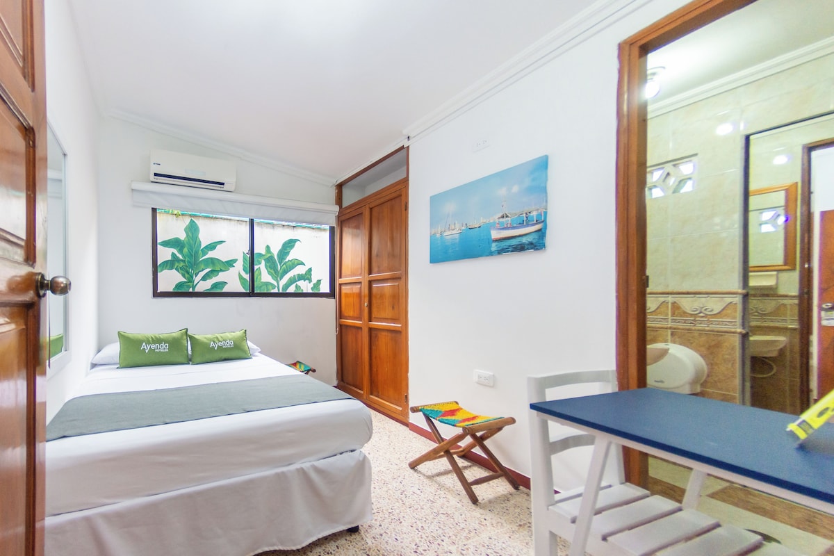 A bedroom with a bed, a desk, and a chair. There is a window with a view of plants and a painting of boats on the wall.