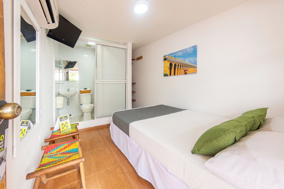 A room with a bed, a green pillow, a colorful woven mat, a painting on the wall, and a sliding door leading to a bathroom.