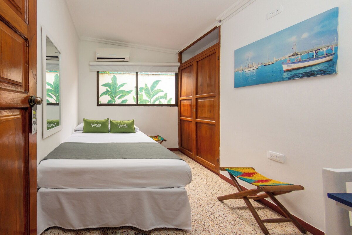 Un dormitorio con cama, ventana con vistas a follaje, equipo de aire acondicionado y un cuadro de un barco en la pared.