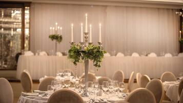 A banquet hall with round tables set for dining, a central candle holder with greenery, and a backdrop of draped curtains.