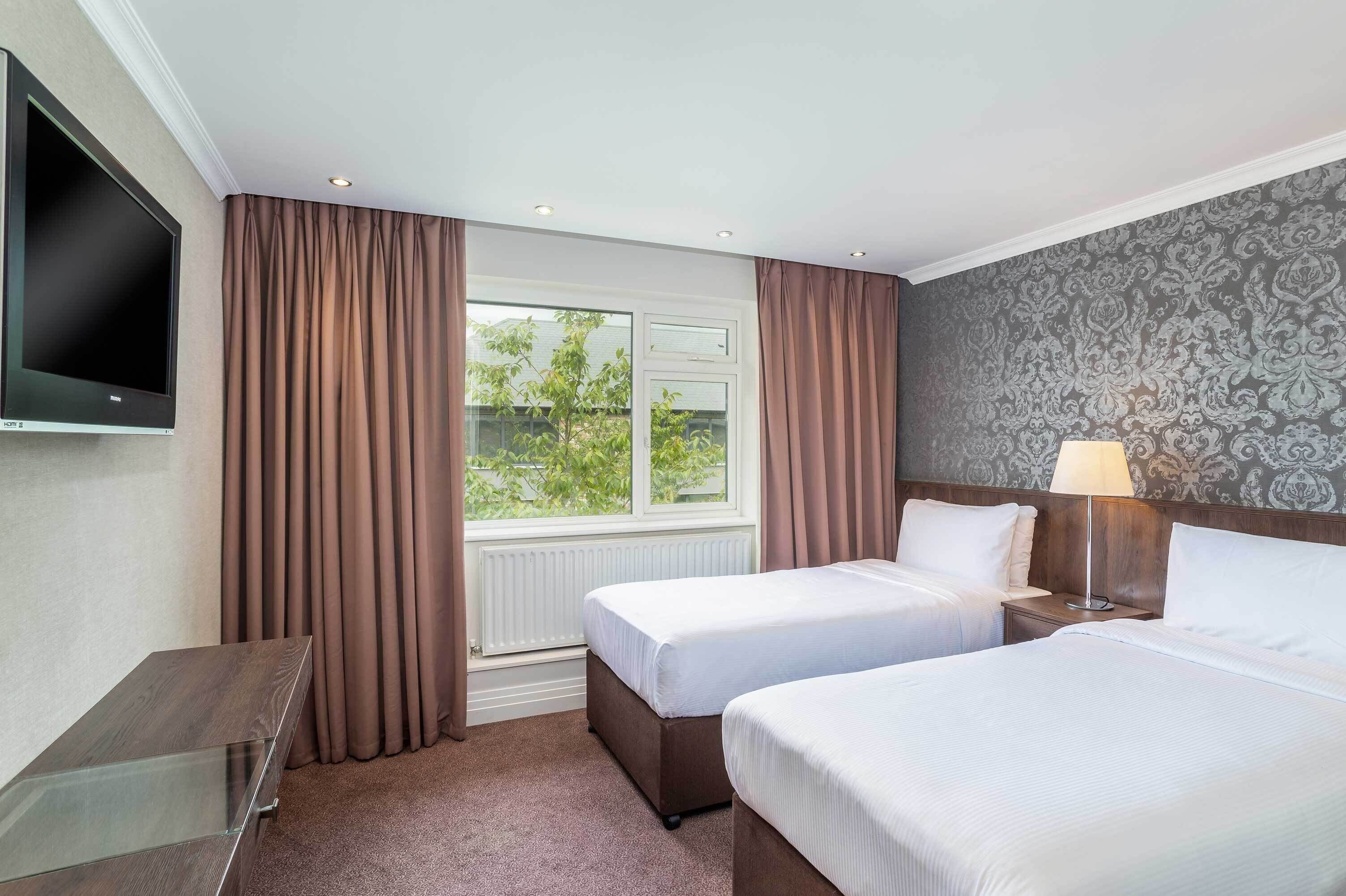 A hotel room with two beds, a TV, a lamp, and a window with curtains.