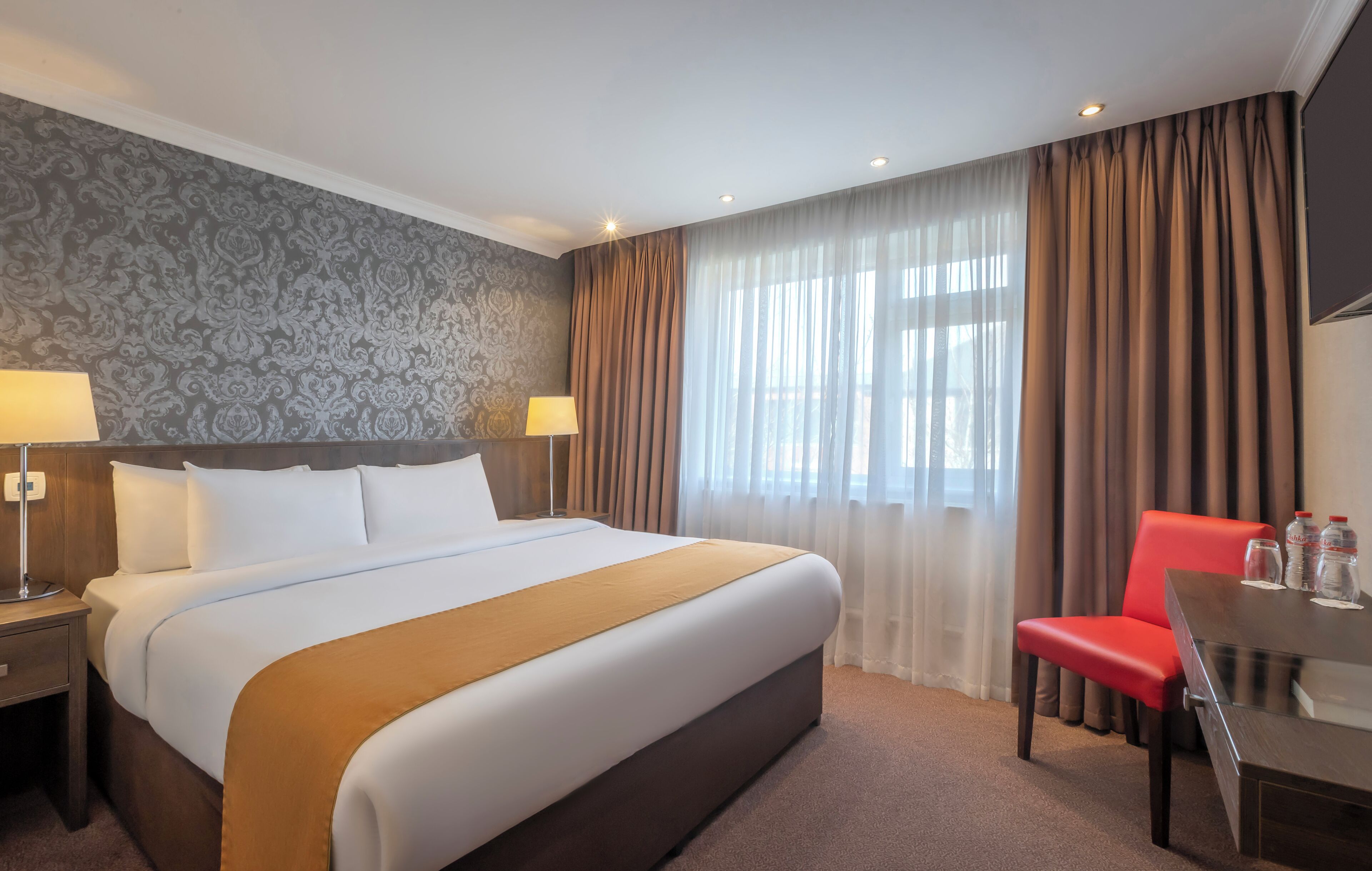 A hotel room with a large bed, a desk, a chair, and a window with curtains.