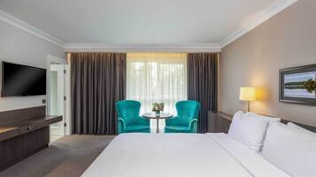 A hotel room with a large bed, a TV, a desk, two chairs, a small table with a vase, and a lamp.