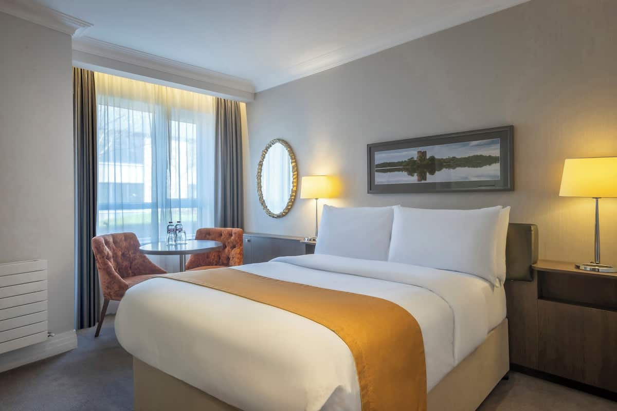 A hotel room with a large bed, a round table, a chair, a mirror, and a painting on the wall.