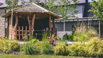 A log cabin structure with a thatched roof, surrounded by a variety of plants and shrubs.