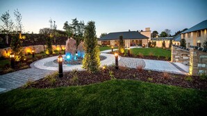 Un jardín ajardinado con una fuente central, senderos de piedra y zonas exteriores bien iluminadas.