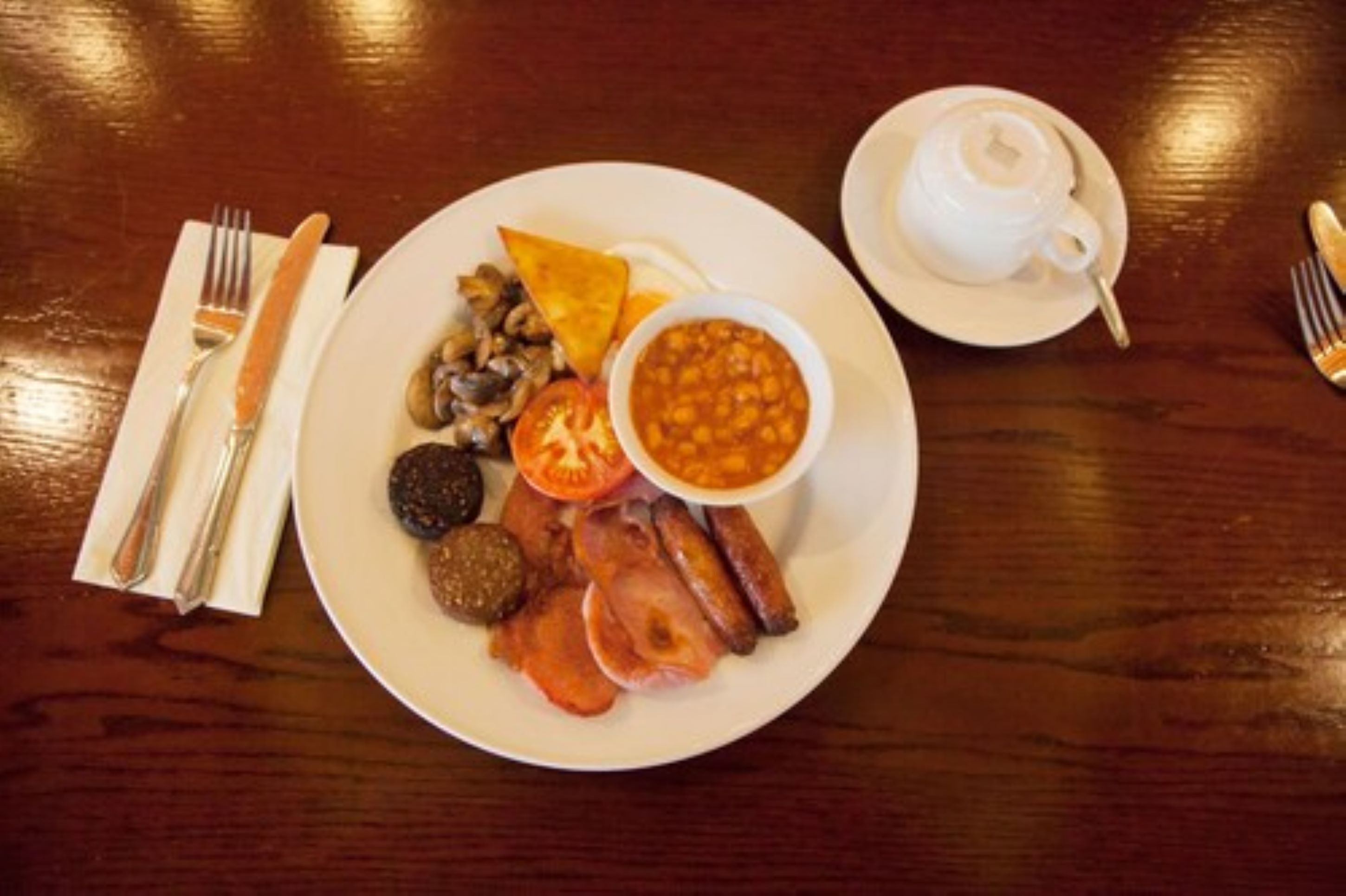 Une assiette de petit-déjeuner comprenant des œufs, du bacon, des champignons, des tomates et des haricots, accompagnée d’une tasse de thé et de couverts, le tout posé sur une table en bois.