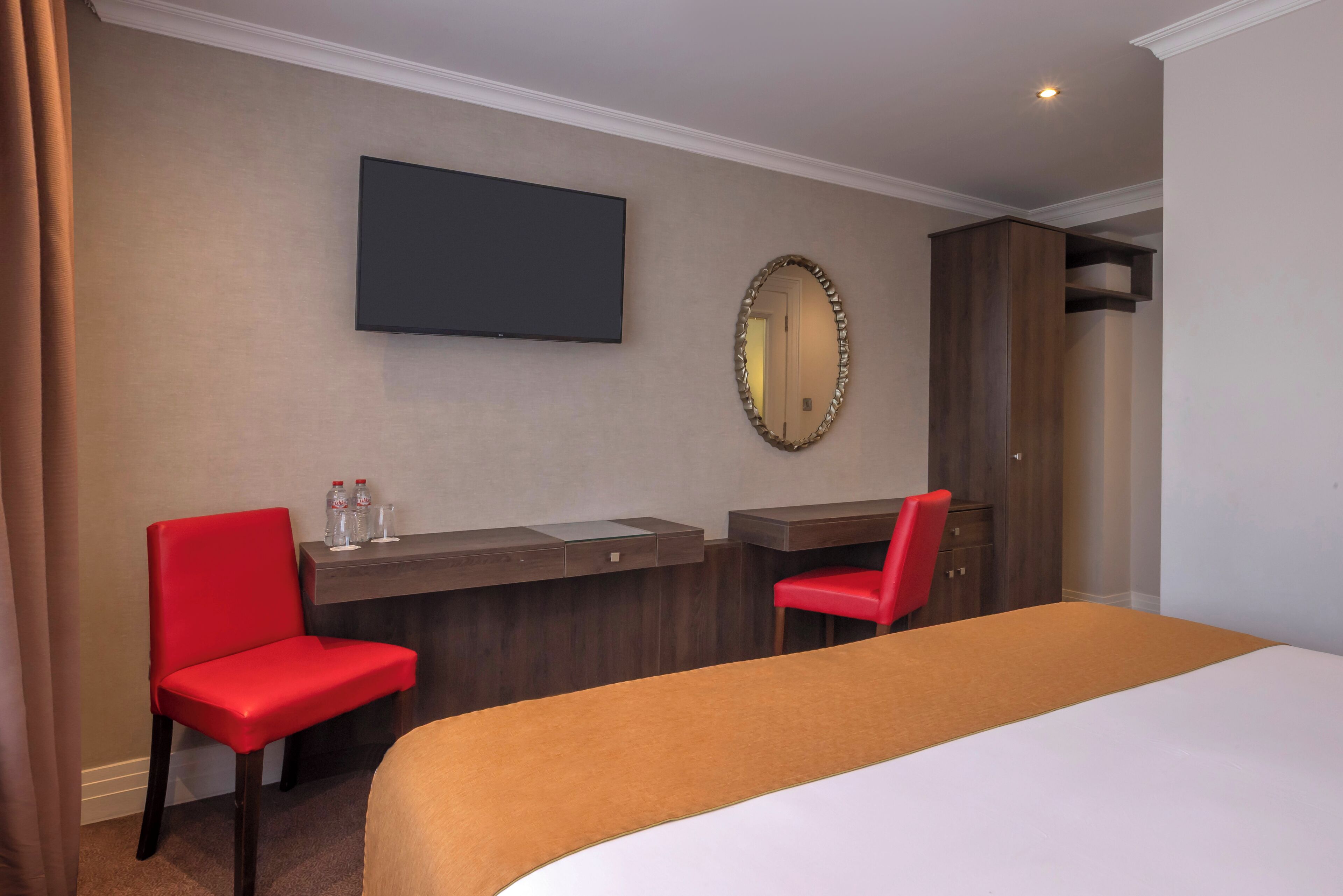 A hotel room with a bed, two red chairs, a desk with a flat-screen TV, a mirror, and a wardrobe.