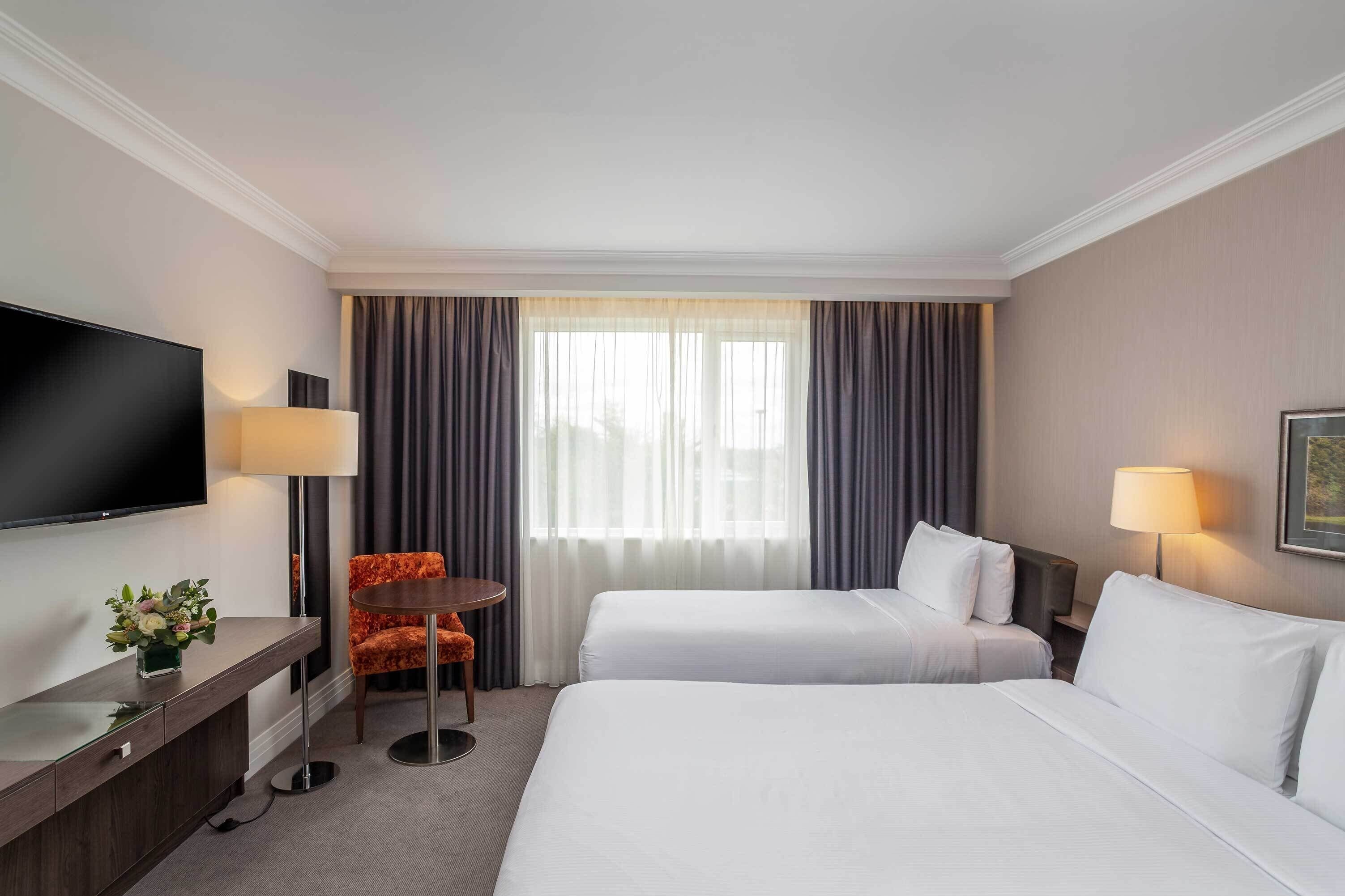 Ein Hotelzimmer mit zwei Betten, einem Fernseher, einem Schreibtisch mit Lampe und einem Sessel.