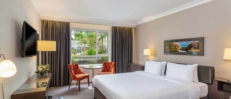 Habitación de hotel con una cama grande, un escritorio con lámpara, un televisor, una mesita con dos sillas y una ventana con cortinas.