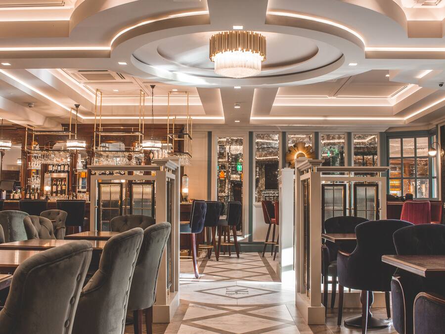 A modern restaurant interior with a central chandelier, multiple dining tables, and a patterned floor.