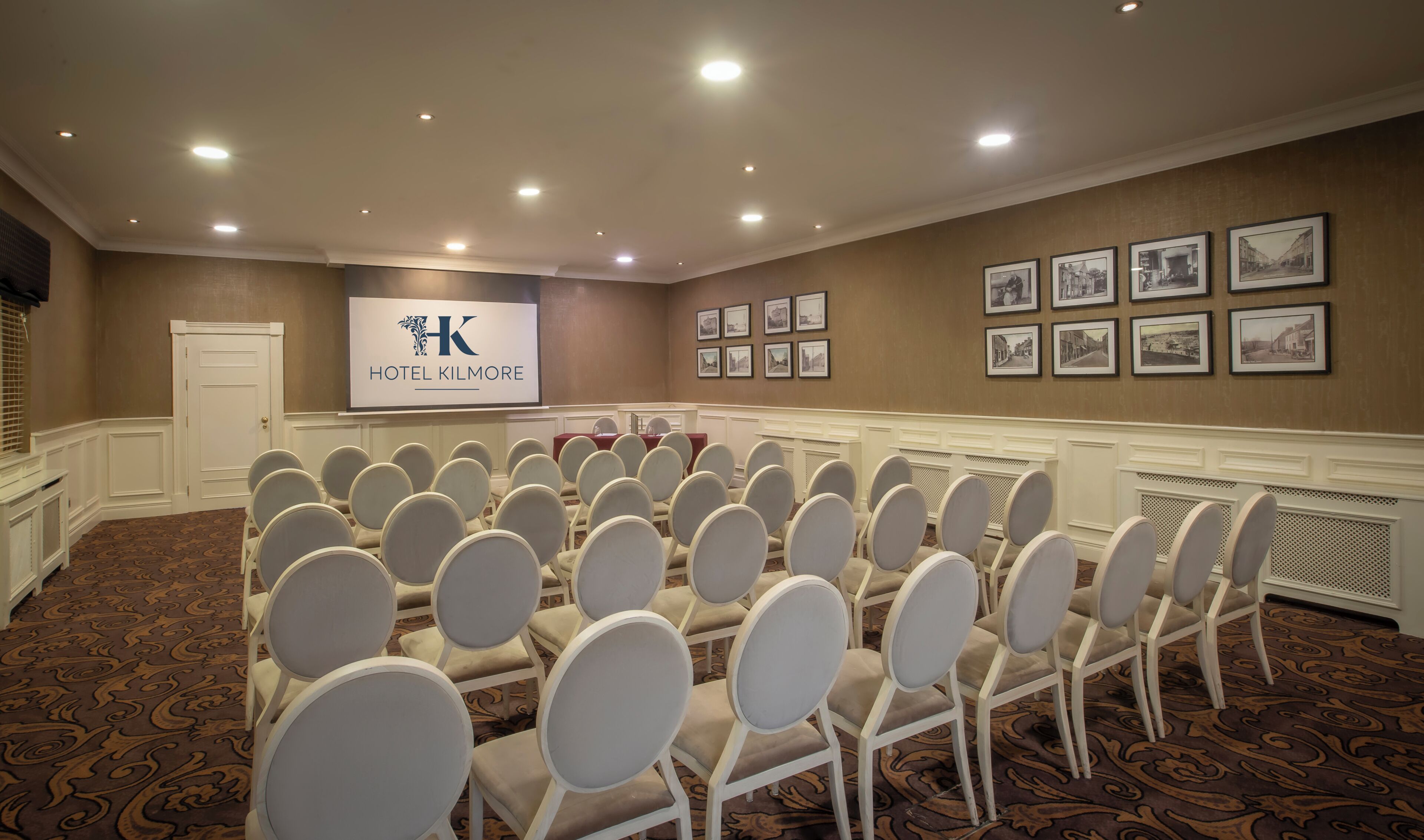 Un salón de conferencias con filas de sillas blancas, un atril y un letrero que dice 'HOTEL KILMORE'.