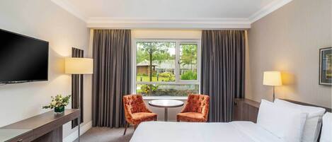A hotel room with a large bed, two armchairs, a small table, a TV, and a window with curtains.