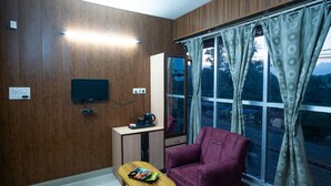 Une pièce aux murs en bois, un téléviseur fixé au mur, une petite table avec un ordinateur portable et des en-cas, un fauteuil violet et une fenêtre avec des rideaux.