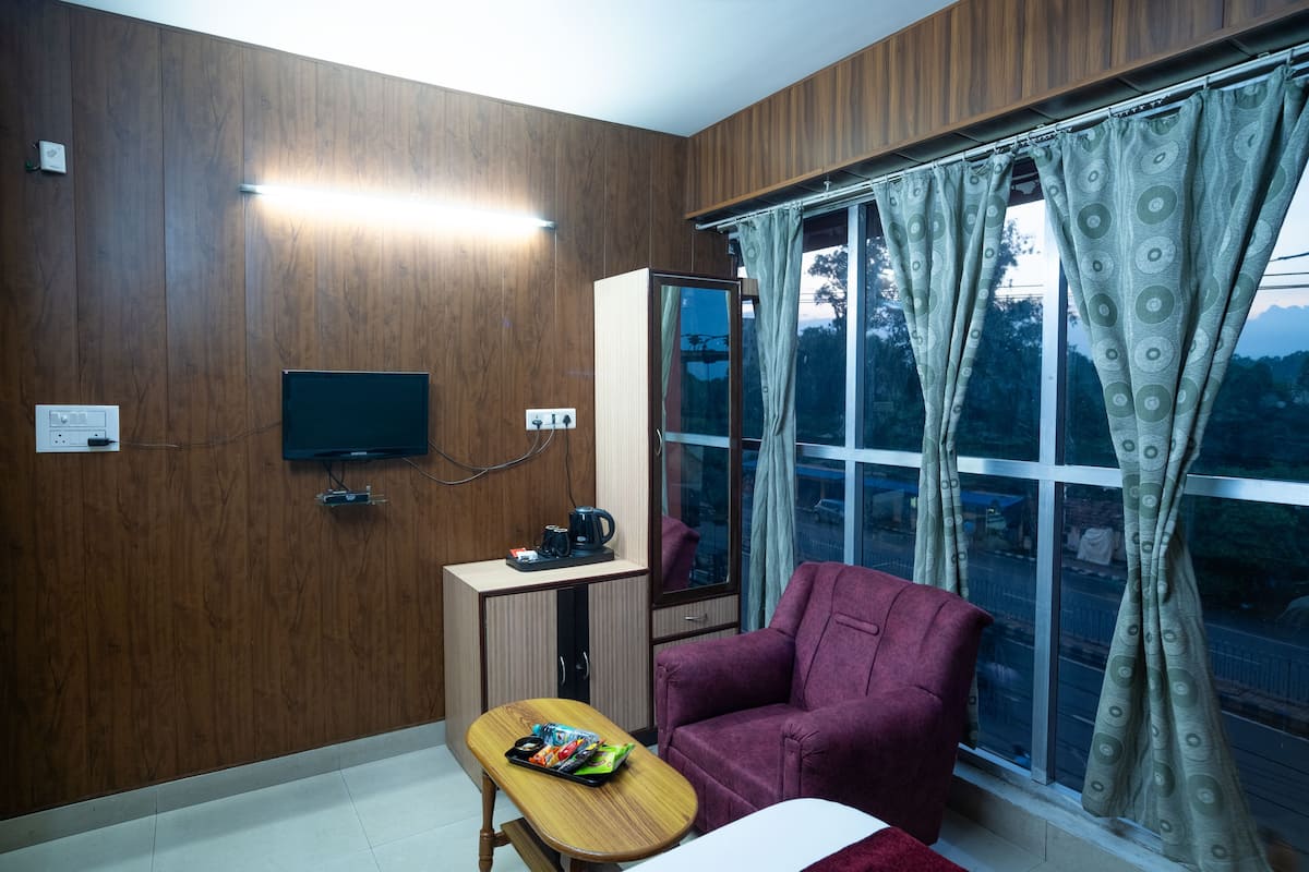 Una habitación con paredes de madera, un televisor montado en la pared, una mesa pequeña con una computadora portátil y aperitivos, un sillón morado y una ventana con cortinas.