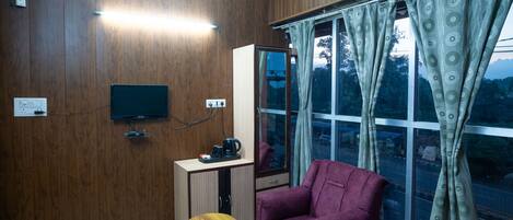 A room with wooden walls, a television mounted on the wall, a small table with a laptop and snacks, a purple chair, and a window with curtains.