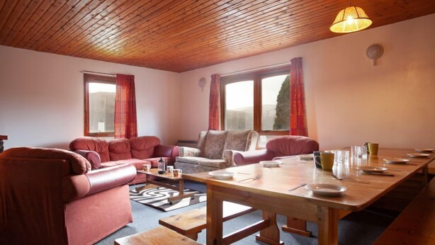 Een gezellige woonkamer met een houten plafond, rode banken, een houten eettafel en grote ramen met gordijnen.