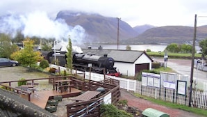 Een model treinset met een stoomlocomotief, een picknickplaats met banken, en een uitzicht op een meer en bergen in de achtergrond.