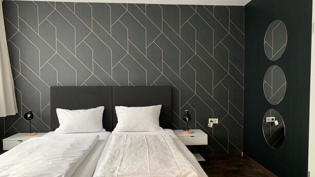 A hotel room with a modern design, featuring a large bed with two white linens, a dark headboard, and a geometric patterned wall. There are two bedside tables with lamps and a mirror on the wall. - MOTELO Bielefeld (Bielefeld)