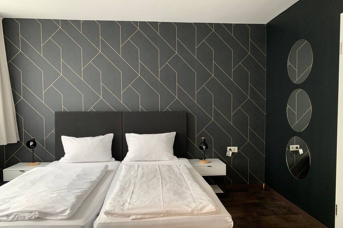 A hotel room with a modern design, featuring a large bed with two white linens, a dark headboard, and a geometric patterned wall. There are two bedside tables with lamps and a mirror on the wall.