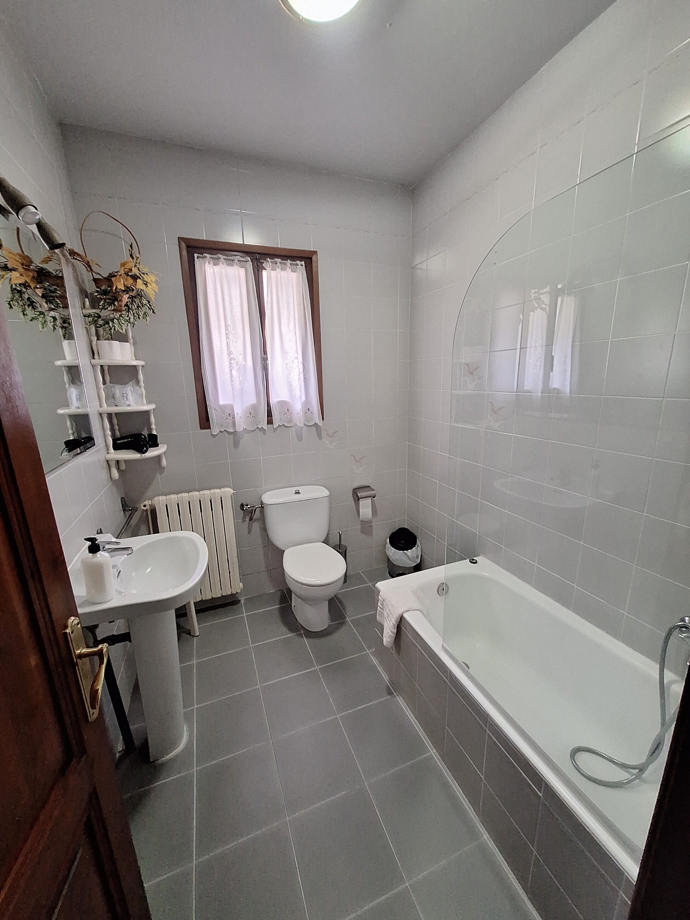 A bathroom with a white sink, toilet, and bathtub. There is a window with curtains, a radiator, and a mirror with shelves above it.