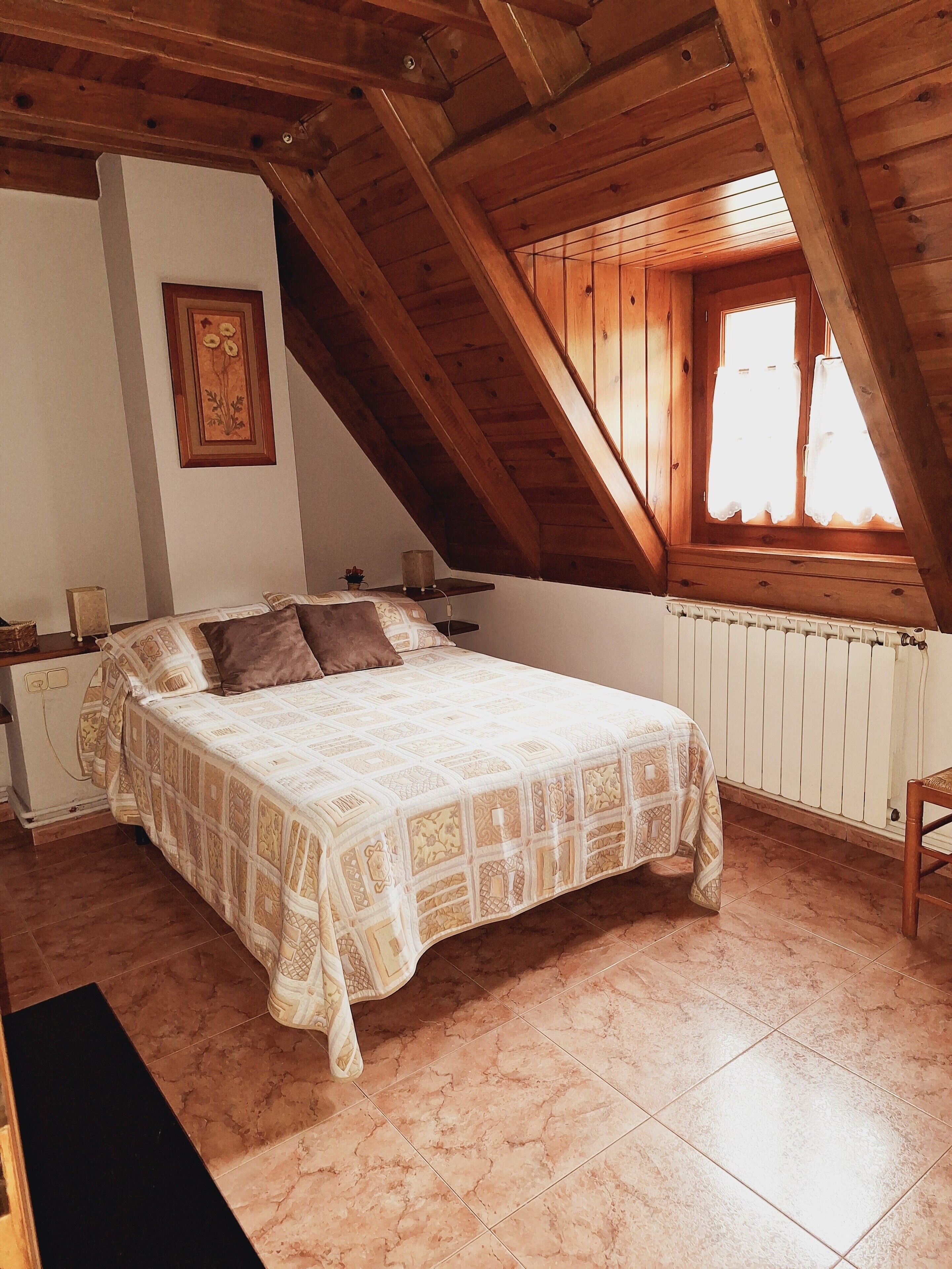 A bedroom with a bed, wooden beams, a radiator, and a window with curtains.