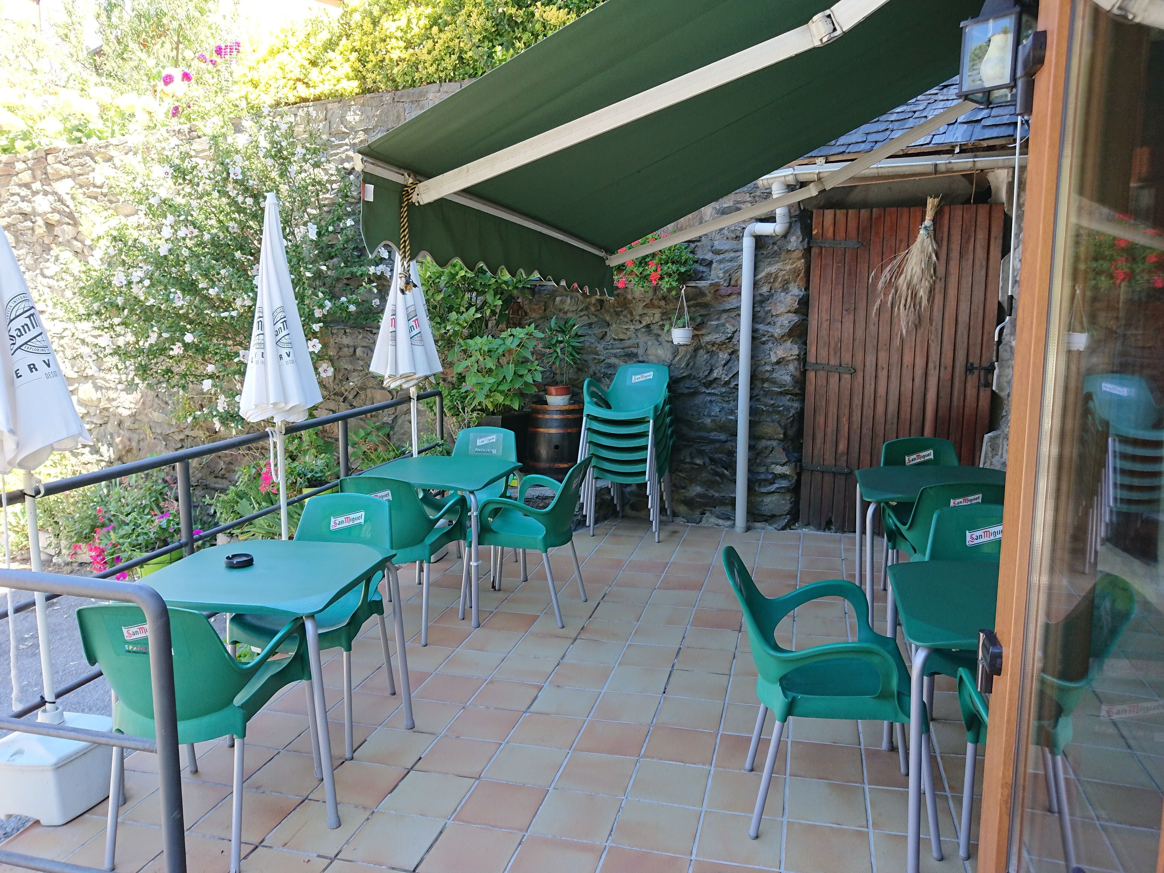Un patio al aire libre con mesas y sillas de plástico verde, un toldo verde y una pared de piedra con plantas.