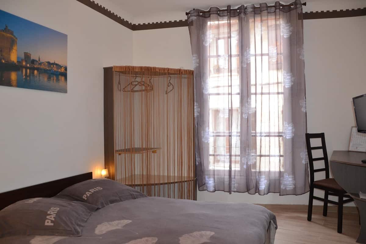 A bedroom with a bed, a desk with a chair, a wardrobe, and a window with sheer curtains.
