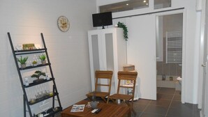 A living room with a wooden table, two wooden chairs, a black metal shelf with plants, a clock, and a television on a stand. - B&B La Maison d'Olivier (Beaucaire)