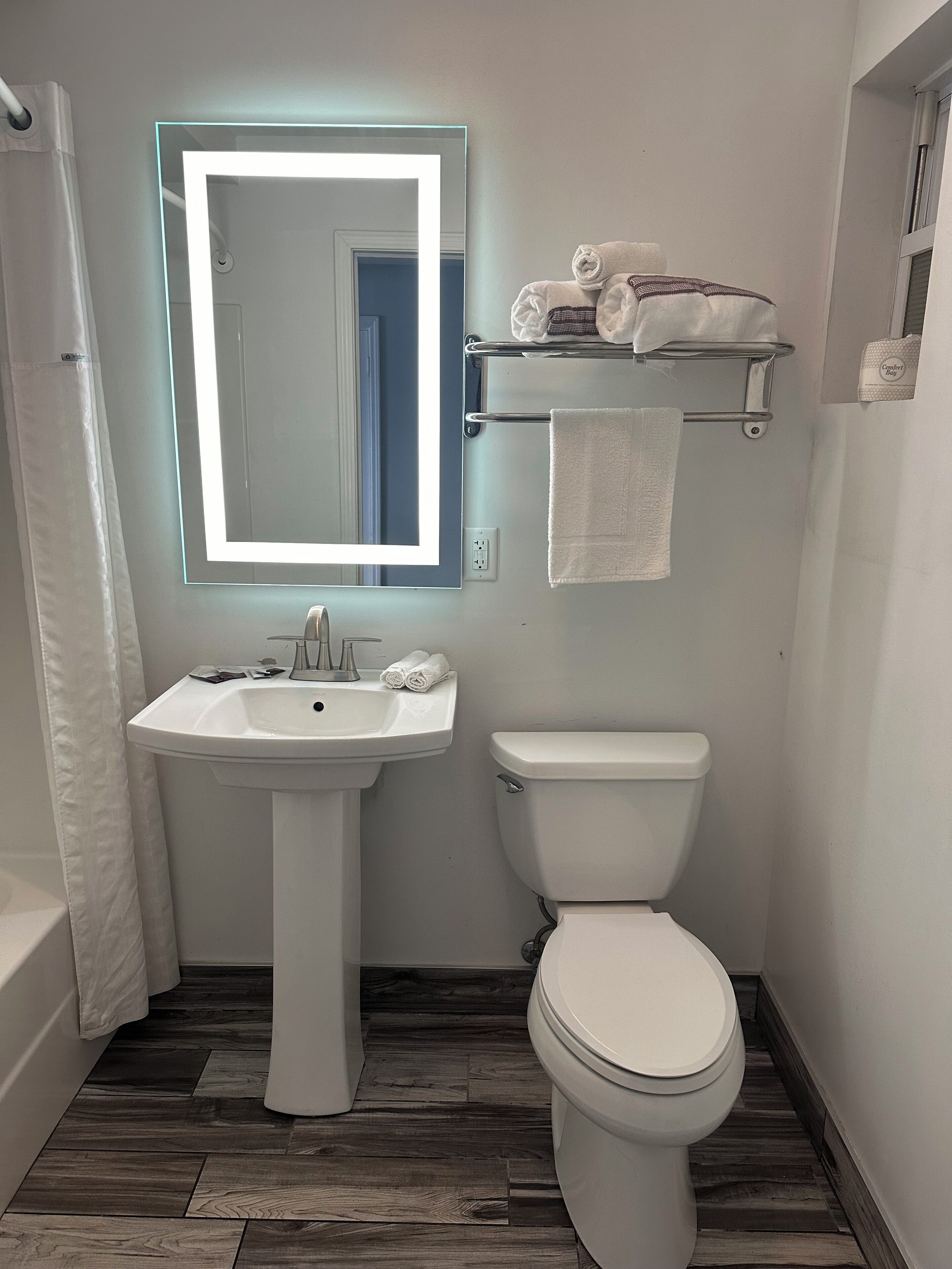 A bathroom with a white sink, a toilet, and a mirror with a light. There are towels on a rack and a shower curtain.
